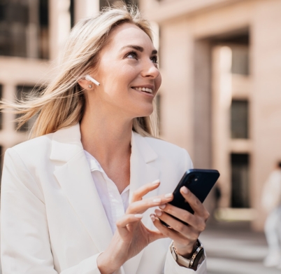 Excited young blonde businesswoman woman making video call by phone, earphones, with happy facial expression smiling goes outdoors against building. Pretty lawyer typing message. Happy cute student